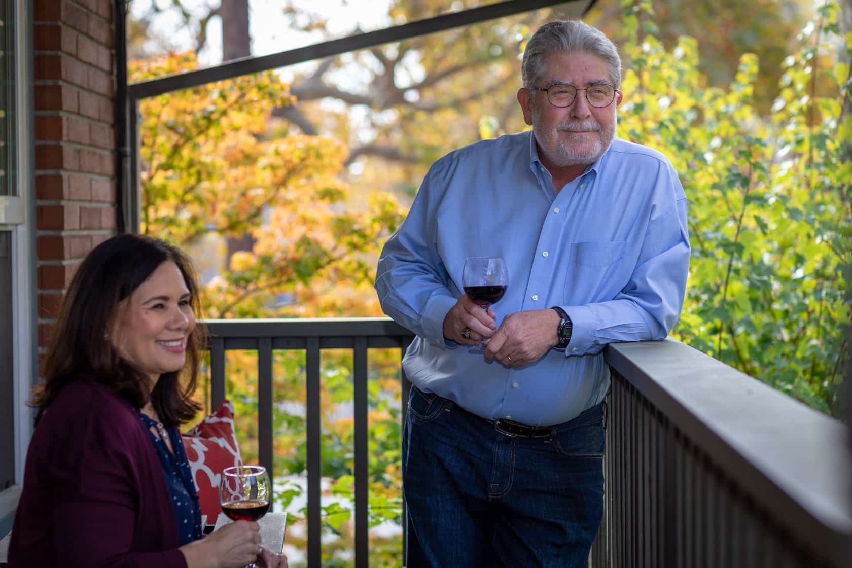 Older gentleman and gentlewoman both with a glass of wine leaning against a railing outside looking relaxed